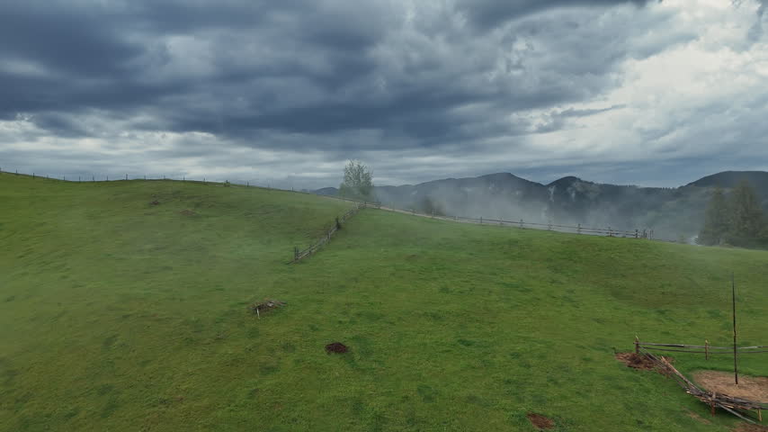 Stormy mountain meadow under dark dramatic clouds with mist drifting through distant hills, Green alpine pasture and wooden fences create a moody rural landscape.