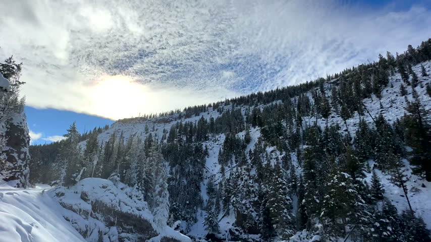 A Scanning Shot Down from a Beautiful Blue Sky Through a Snow Covered Forest of Pine Trees to Reveal a Waterfall and River Rushing Through the Snow Covered Landscape
