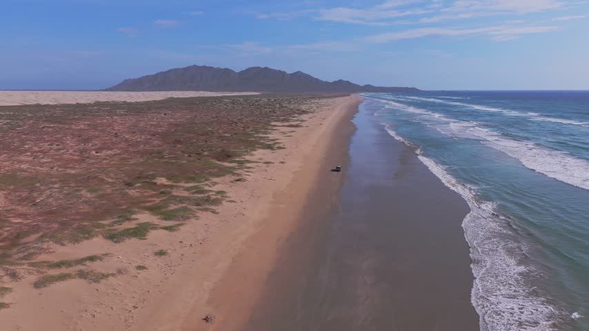 Drone view tracking a truck driving along the wet shoreline of a vast deserted beach with dunes and mountains behind.