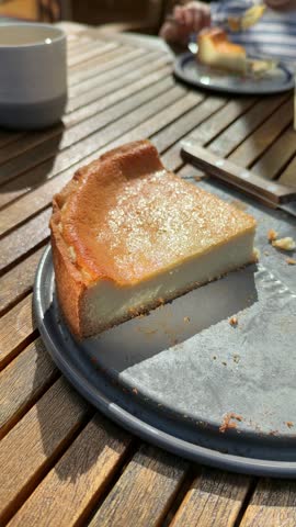 Close-up vertical video of a large slice of golden baked cheesecake on a grey plate on a sunny wooden garden table, with a coffee cup and a person eating cake in the background.