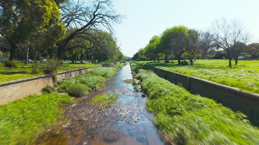 A dynamic, fast-moving drone shot races low along a winding canal through a lush green park, weaving between overhanging tree branches and past palm trees, grassy lawns, and a concrete retaining wall.