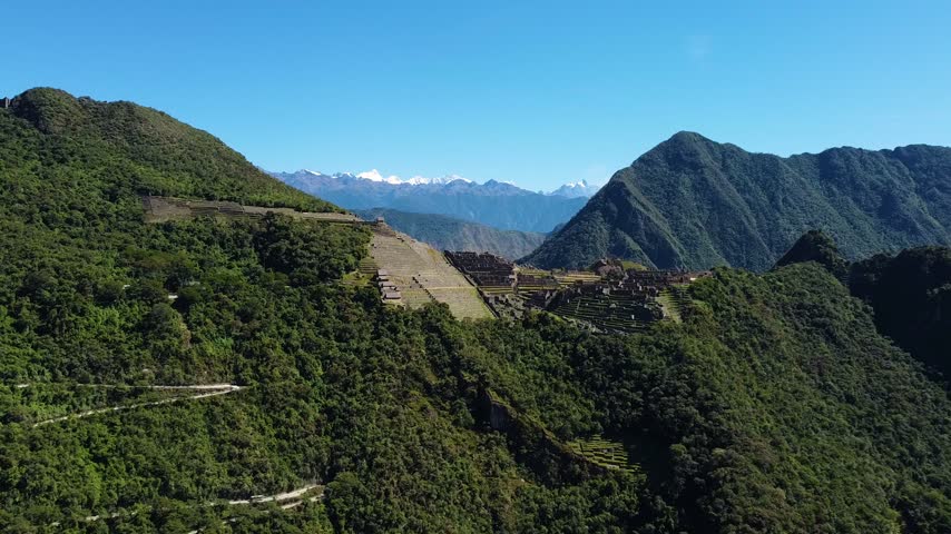 Aerial view of the Historic Sanctuary of Machu Picchu nestled among lush green Andean mountains under a clear blue sky in Urubamba Province, Cusco, Peru.