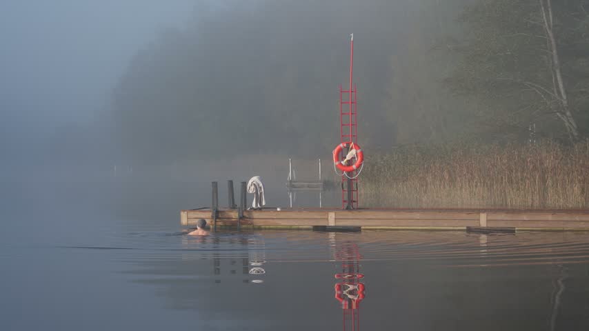 Middle aged man entering cold lake water from wooden dock early autumn morning foggy landscape Finland