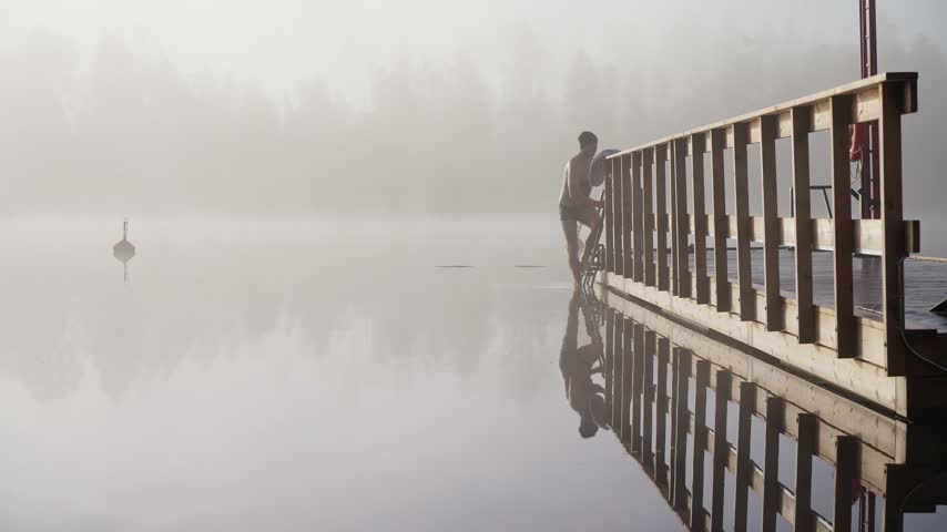 Middle aged man entering cold lake water from wooden dock early autumn morning foggy landscape Finland