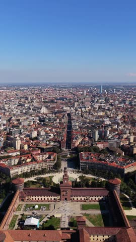 Vertical forward flight over Castello Sforzesco courtyard toward Filarete Tower and Piazza Castello fountain in Milan, Italy, Milan Cathedral in the distance under blue sky