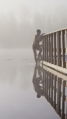 Middle aged man entering cold lake water from wooden dock early autumn morning foggy landscape Finland