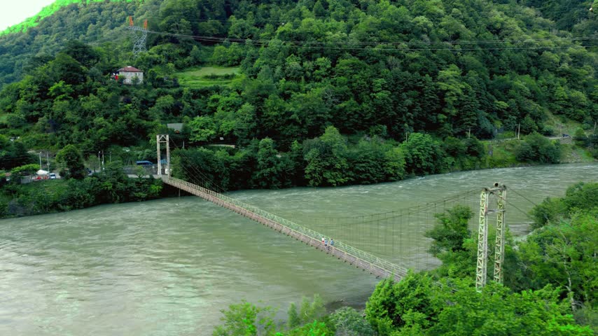 Lush green mountain slopes surround a wide, fast-flowing river with a long pedestrian suspension bridge connecting the banks, creating Georgia
