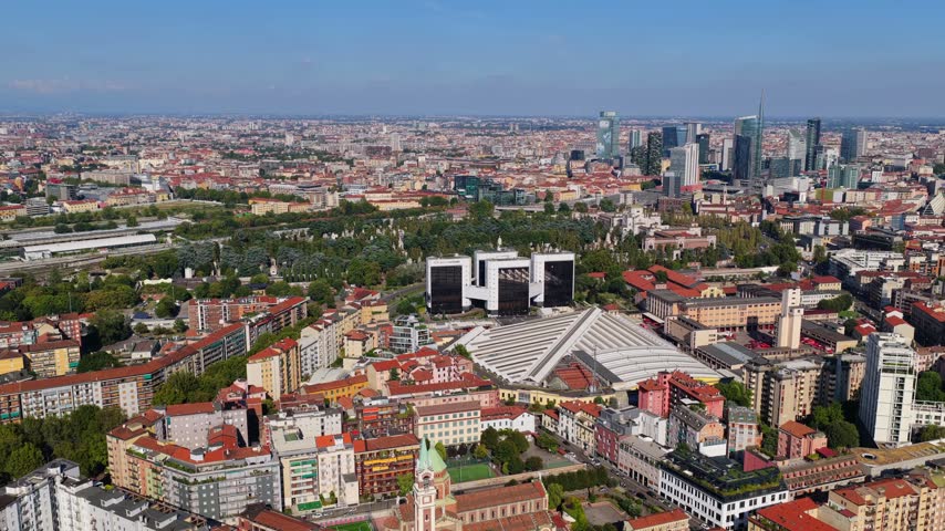 Wide aerial panorama over red rooftops, a white roofed hall, Cimitero Monumentale and railway yard toward the Porta Nuova skyline and UniCredit Tower, sunny day in Milan, Italy
