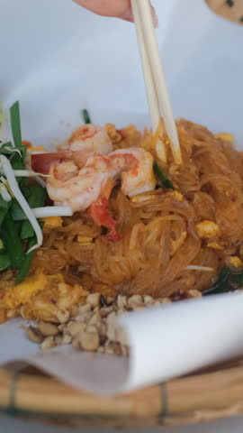 Vertical video. Close-up of a woman using chopsticks to eat traditional Pad Thai noodles with seafood from a bamboo plate