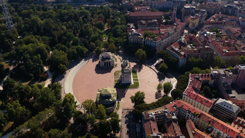 High angle orbit around Arco della Pace and its twin toll houses on round Piazza Sempione in Milan, Italy, beside Parco Sempione trees on a sunny day