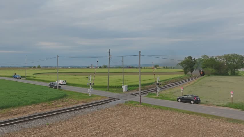 Aerial view of a steam train approaching a rural railroad crossing with cars waiting at the gates amidst green fields in Ronks, Pennsylvania, United States.