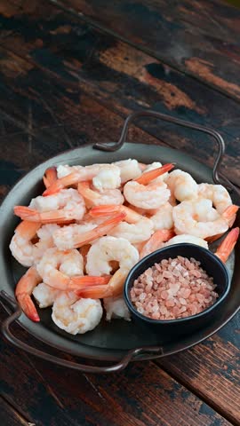 Vertical view of cooked peeled shrimp and coarse pink seasoning rotating in a dark serving dish on a rustic wooden tabletop.