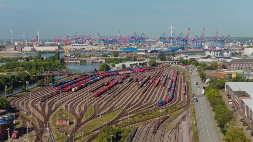 Aerial view of a freight rail yard backed by towering container cranes and a container ship in Hamburg, Germany, pulling back to reveal a canal and truss bridge