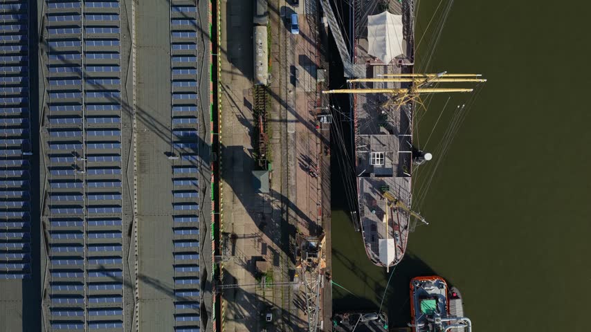 Top down view flying along the historic four-masted barque Peking from stern to bow, yellow masts and yards over dark harbor water beside a quay in Hamburg, Germany
