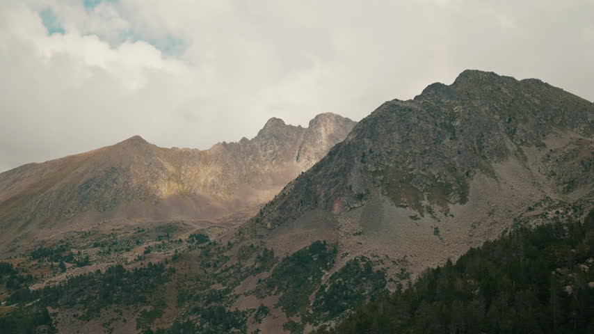 Majestic Pyrenean mountain peaks in Principality of Andorra, with Pinus uncinata pines, sunlight and cloudy sky backdrop. Scenic view of rocky mountain ranges at moody cloudy weather and sunlight play