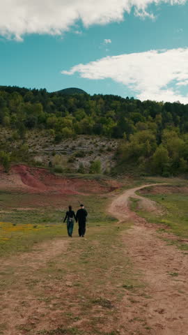Two people walking on dirt path at Congost de Mont-rebei, Spain. Couple hiking towards green forest hill under blue cloudy sky. Red soil and yellow flowers meadow scene, moving forvards along road