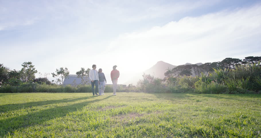 Diverse friends walking across field in red, green, light jacket, reaching front and posing shadows. Backlit, silhouettes, trio, sunlit, meadow, nature, lensflare