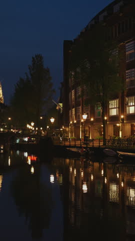 Illuminated canal with street lights and Montelbaanstoren tower reflection at night in Amsterdam, Netherlands. Night view of canal with warm lantern reflections on calm water and historic bridge