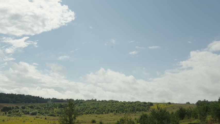 Fluffy white clouds drift slowly across a bright blue sky above a scenic rolling green countryside landscape with young trees and open fields.