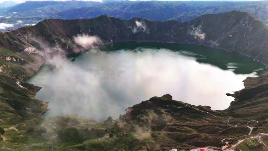 Aerial view of the turquoise Quilotoa Lake inside a volcanic crater surrounded by rugged mountains and clouds in Cotopaxi, Ecuador.