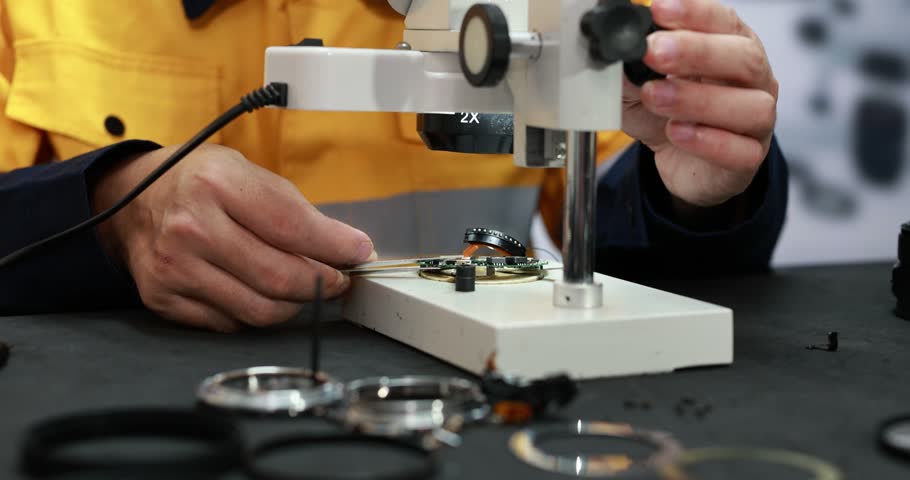 Watchmaker in a yellow work coat repairs a camera lens under a precision magnifier, with small tools and lens parts on a dark workbench.