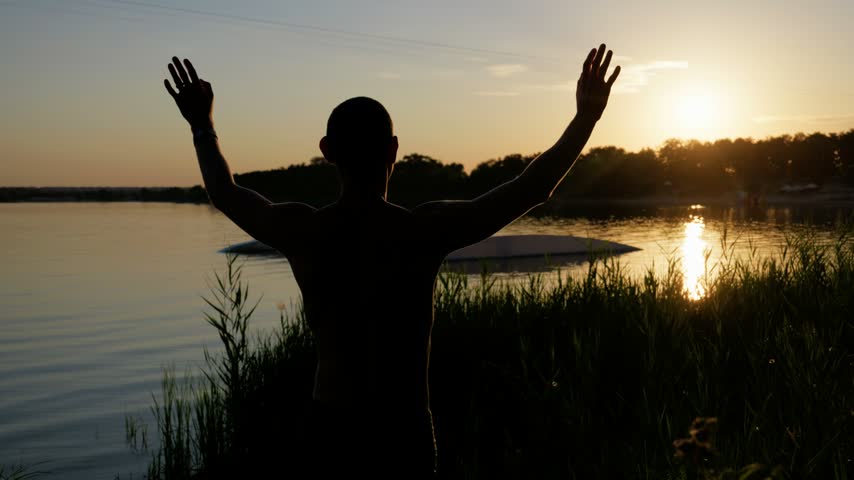 Rear silhouette of an athletic man standing by tranquil lake reeds with outstretched arms during golden hour sunset. Warm evening light reflects on calm water along tree-lined shore.