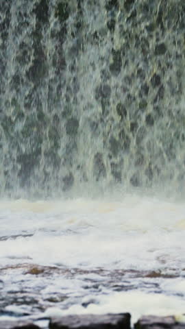 Close-up view from the lower part of a waterfall, focusing on the falling streams and turbulent water at its base. The impact creates white foam and swirling currents across the surface, while blurred 
rocks appear in the foreground. Gentle slow motion emphasizes the changing texture and flow. Vertical video, natural daylight and a low viewpoint capture the dynamic character of the falls.