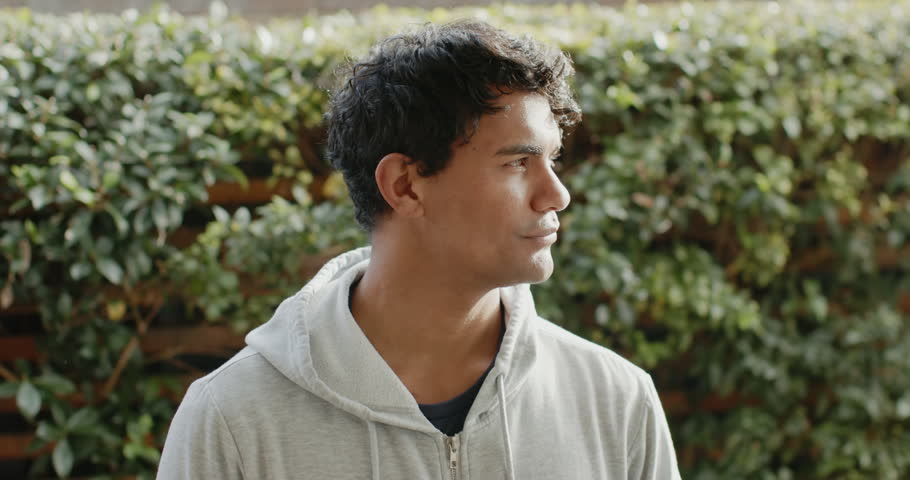 African American man camera-prompted, turning toward lens in yard, wearing gray hoodie, smiling. Portrait, outdoor, garden, hedge, fence, adult, male