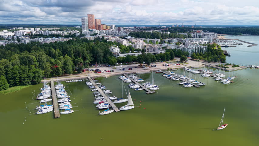 Aerial view of Vuosaari marina in Helsinki, Finland, showing sailboats docked at piers, residential buildings, lush green forest and calm bay waters under cloudy summer sky