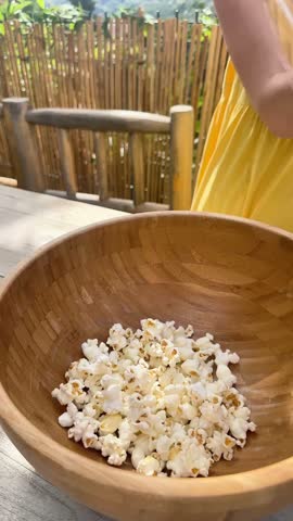 High-angle shot of a person pouring freshly popped popcorn into a large wooden bowl placed on a rustic wooden table outdoors, with a person wearing a yellow dress standing nearby.