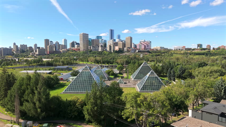 Forward aerial passes above the glass pyramids and greenhouses of Muttart Conservatory with downtown Edmonton rising behind on a bright summer day.