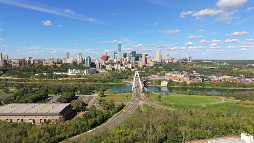 Forward aerial flight follows Walterdale Bridge across the North Saskatchewan River toward downtown Edmonton in bright summer sunshine beneath a clear blue sky.