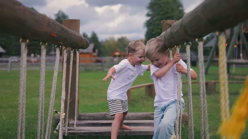 Two boys engage in playful activity on a rope bridge, balancing and laughing together in a park setting surrounded by greenery and trees