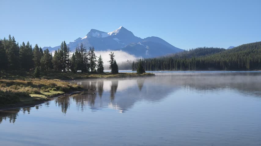 Maligne Lake reflects snow-capped mountains and green evergreen trees under a clear blue sky, with mist rising from the calm water in Jasper National Park, Alberta, Canada