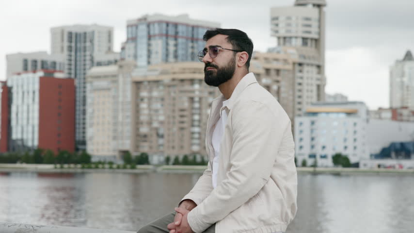Young bearded man in three-quarter profile seated on waterfront railing with folded hands, beige jacket and round tinted sunglasses, overcast city skyline bokeh background