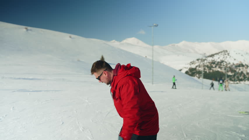 A man in a red jacket is seen snowboarding down a sunny, snowy mountain slope. He enjoys the winter sport with a beautiful mountain landscape in the background.