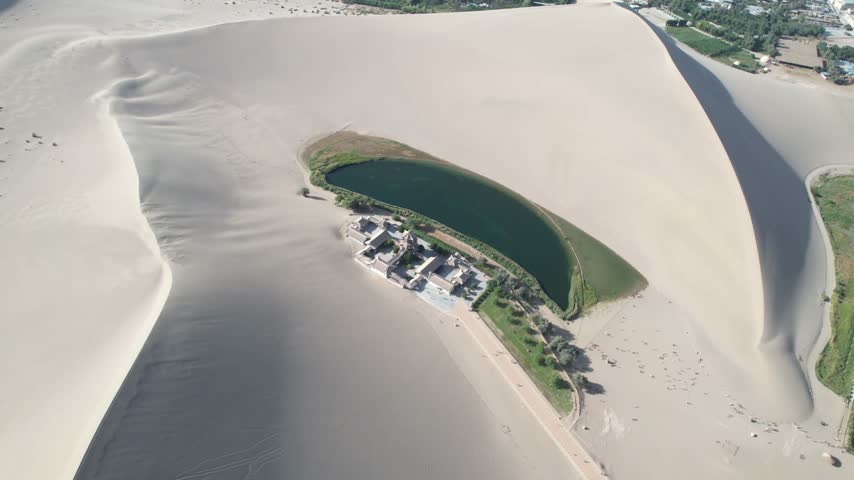 Aerial view of Crescent Lake oasis in Dunhuang, China. Drone circles above the crescent-shaped water body surrounded by vast sand dunes and traditional pagoda-style buildings on a sunny day.