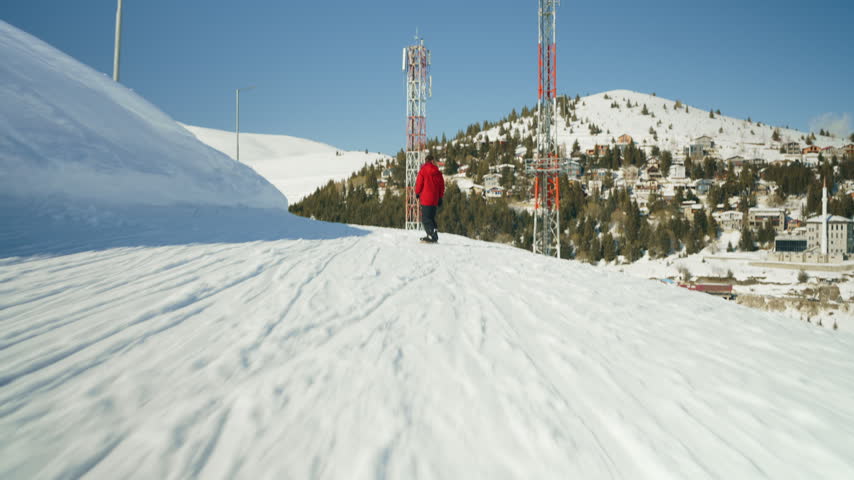 A man in a red jacket stands on a snowy mountain ridge, with two communication towers in the background. The winter landscape features a clear blue sky, pristine snow, and a distant village.