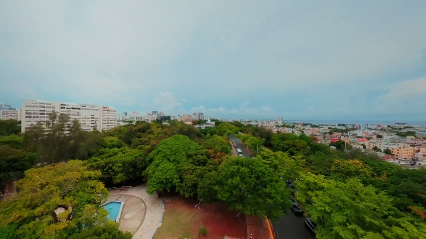 Aerial view of Mirador Sur park with lush green trees, a winding road with traffic, and the city skyline under a cloudy sky in Santo Domingo, Distrito Nacional, Dominican Republic.