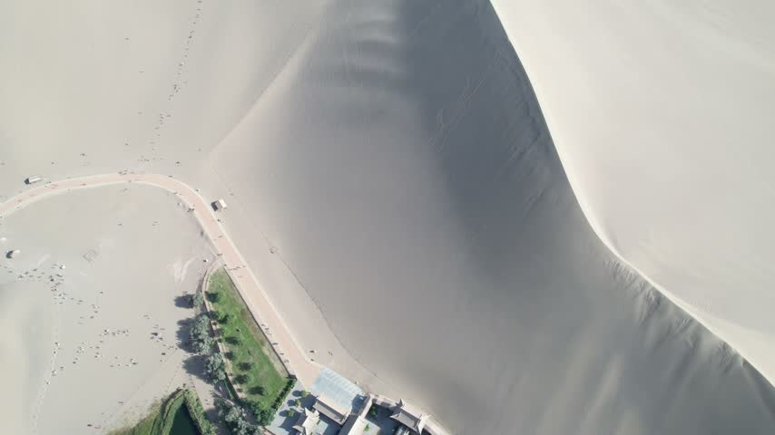 Aerial top-down view of Crescent Lake oasis surrounded by the Singing Sand Dunes near Dunhuang, Gansu, China. The drone shot reveals the crescent-shaped lake, traditional architecture, and vast desert