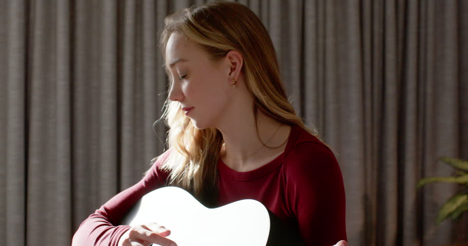 Placing pick on strings, adult woman practicing strumming guitar at home in maroon top, jeans. Musician, acoustic, fretboard, chords, fingers, sunlit, cozy