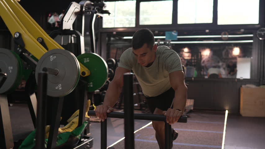 Strong muscular man intensely pushing a weighted sled on a track inside a modern gym, engaging in a powerful strength and conditioning workout for building endurance and functional fitness