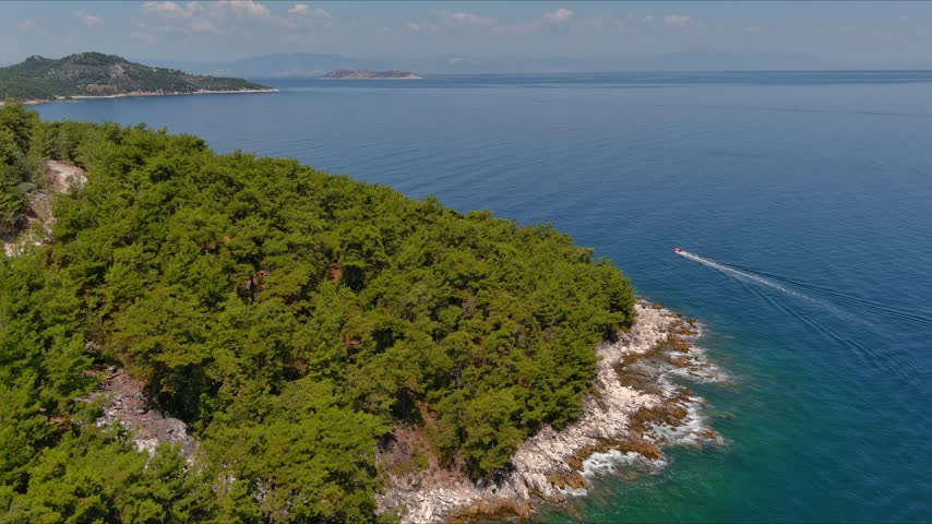 Aerial view of the pine-covered coastline and rocky shore of Thasos with a small boat navigating the blue Aegean Sea in Thasos, Greece.