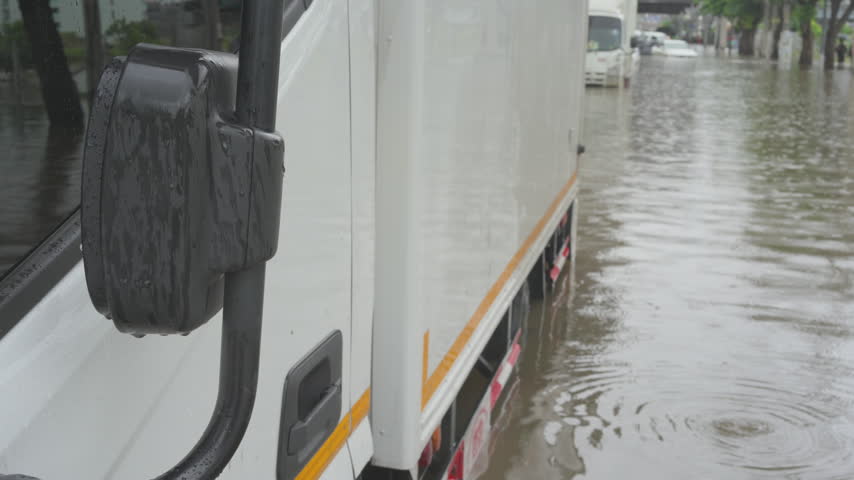 A commercial vehicle navigates a submerged urban road during a severe weather event with other traffic visible in the distance through the rain