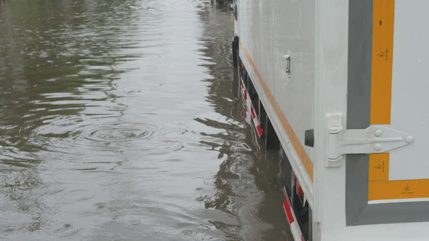 A commercial vehicle navigates a submerged urban road during a severe weather event with other traffic visible in the distance through the rain