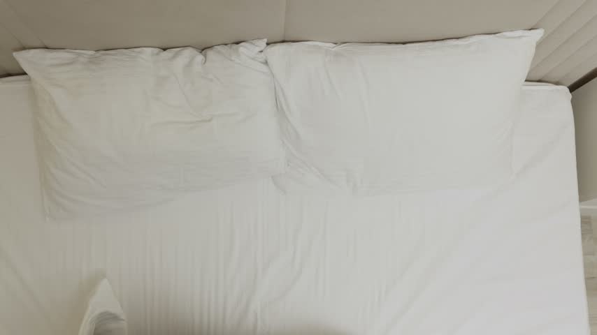 Man, woman lying together on white bedding from above, man embracing woman from behind in tender overhead shot. Soft natural lighting, peaceful intimate moment between partners in bedroom setting.
