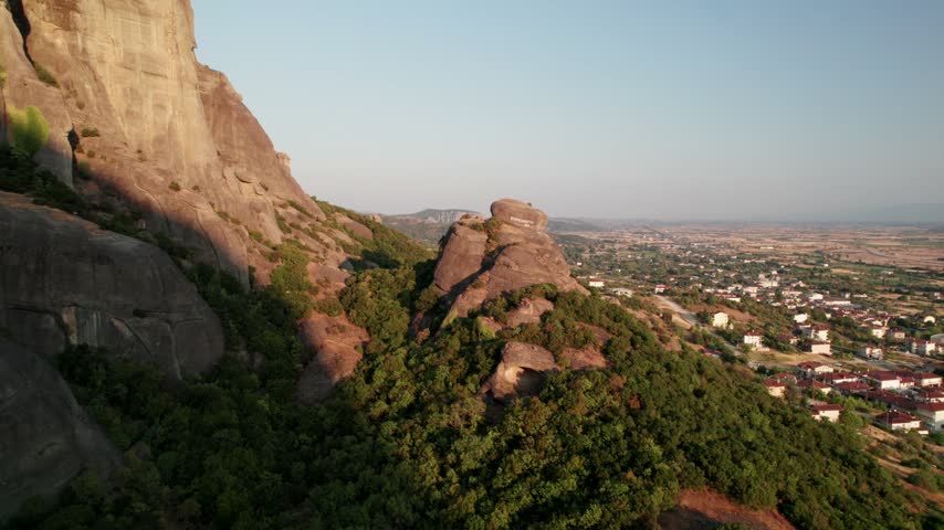 Aerial view of Meteora rock formations with lush green vegetation and the town of Trikala in the background under a clear sky in Trikala, Greece.