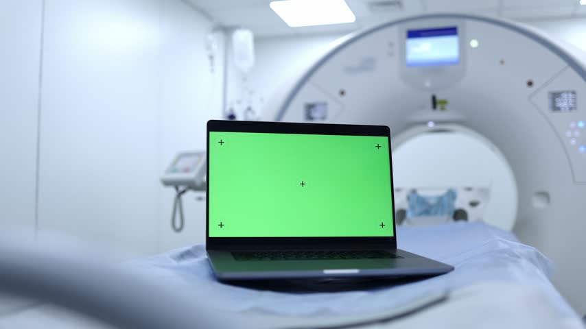 Laptop with chroma key screen on a diagnostic table in front of a CT scanner in a modern medical clinic.