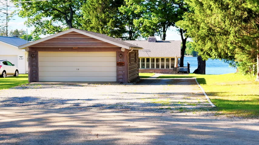White travel trailer sits at a shaded wooded campsite surrounded by mature trees and summer greenery.