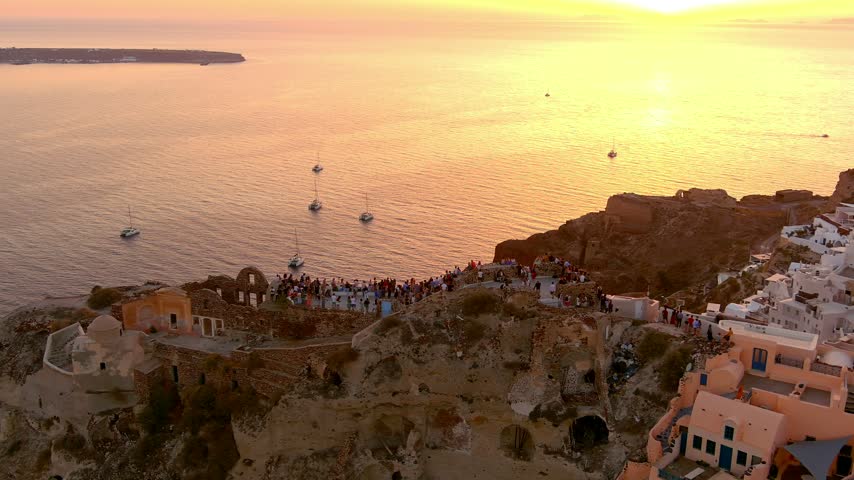 Aerial view of the sunset over the cliffs and white buildings of Oia with sailboats in the Aegean Sea in Islands, Greece.